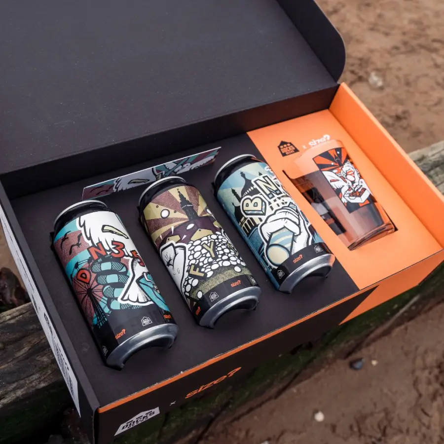 Four Docks Beers cans and a branded glass in a nice orange and black box laid on the sand
