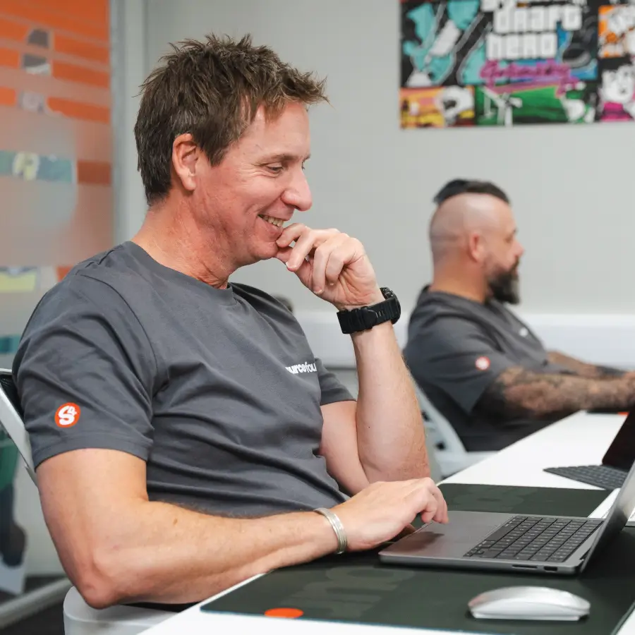 Steve, a Sourcefour employee, smiling whilst working on a laptop, with Alex in the background